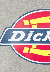 Logo parcial con letras blancas en negrita sobre un banner azul, sobre un fondo circular rojo y amarillo, todo en una tela gris.