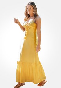Mouwloze gele maxi-jurk met een strikdetail op de borst en een gekruide zoom. De stof heeft een gladde textuur. G gedragen met bruine sandalen.