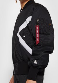 Personne portant une veste bomber noire avec des bandes diagonales blanches, une poche zippée sur le bras avec une étiquette rouge "Remove Before Flight", et un patch casque sur le poignet.