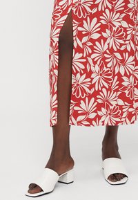 Rotes Maxikleid mit weißen Mustern und Schlitz an der Seite, kombiniert mit weißen Blockabsatzsandalen. Strukturierter Stoff, lockere Passform.