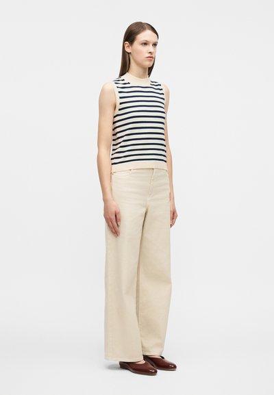 Γυναίκα που στέκεται, φοράει μπεζ αμάνικη μπλούζα με ναυτικούς ρίγες, φαρδιά κρεμ παντελόνια και σκούρα καφέ φλατ παπούτσια σε λευκό φόντο.