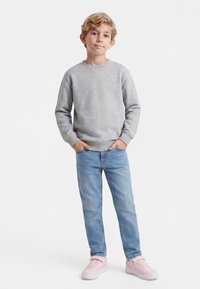 Blond poiss seisab käed taskutes, seljas hall pusa, helesinised teksad ja roosad tossud valgel taustal.