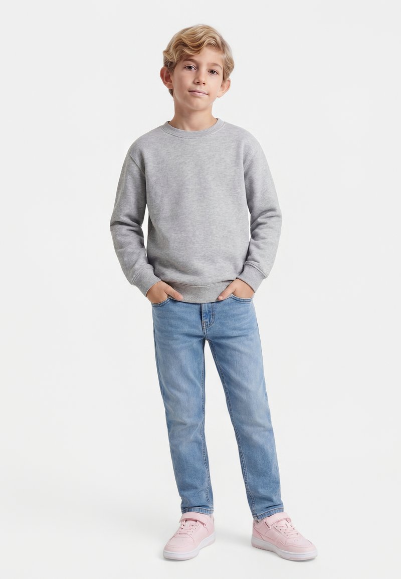 Blond poiss seisab käed taskutes, seljas hall pusa, helesinised teksad ja roosad tossud valgel taustal.