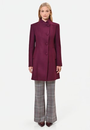 Donna con cappotto viola scuro abbottonato, pantaloni a gamba larga a quadri e scarpe scure con punta, in piedi su uno sfondo bianco semplice.