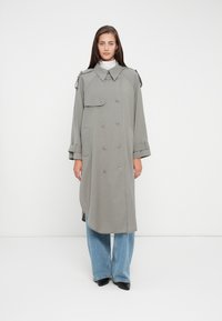 Cappotto lungo grigio con bottoni doppi, spalline e orlo curvo, indossato sopra jeans a gamba larga blu.