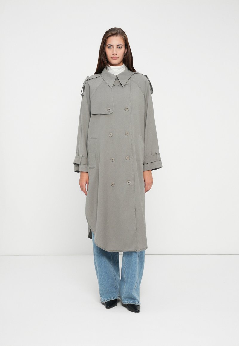 Cappotto lungo grigio con bottoni doppi, spalline e orlo curvo, indossato sopra jeans a gamba larga blu.