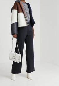 Farbenblockierte Jacke mit braunen, weißen und marineblauen Abschnitten; gemusterte Bluse; weit geschnittene schwarze Hose; weiße Stiefeletten; kleine weiße Handtasche.