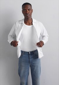 Camisa blanca de botones, de mangas cortas, abierta al frente sobre una camiseta blanca ajustada. Se lleva con unos jeans de mezclilla azul, ligeramente deslavados.