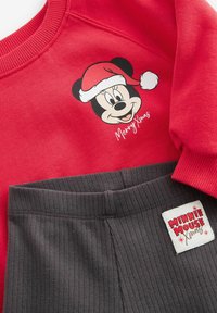 Roter Kapuzenpullover mit einem Mickey-Mouse-Motiv in einem Weihnachtsmannhut und dem Text "Frohe Weihnachten", kombiniert mit grauen Ripp-Hosen und einem "Minnie Mouse"-Label.