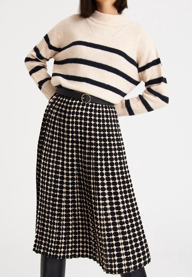 Femme portant un pull crème à rayures noires et une jupe plissée noire et crème à pois avec une ceinture noire sur fond blanc.