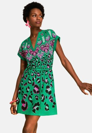 Kobieta w zielonej sukience z krótkim rękawem z różowo-czarnymi wzorami w cętki i serca, uzupełniona o różowe kolczyki w kształcie kwiatów i bransoletki.