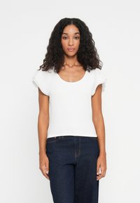 Femme aux cheveux bouclés et foncés portant un haut blanc à manches courtes et un jean bleu marine, se tenant devant un fond blanc uni.