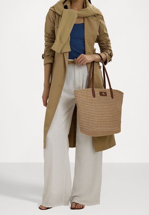 Torebka z plecionego słomy z brązowymi skórzanymi uchwytami, prostokątny kształt i naszywka z logo marki. Modelka nosi lekką trenczową kurtkę oraz spodnie o szerokich nogawkach.
