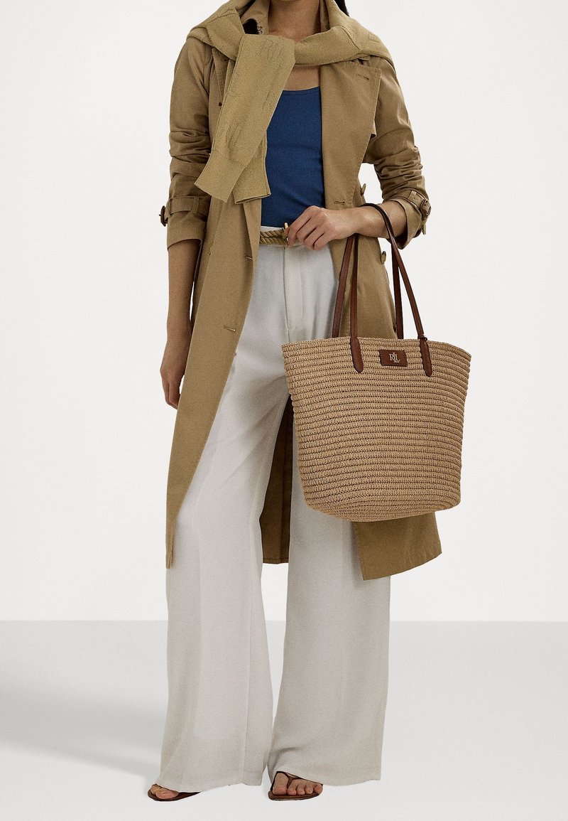 Tkaná slaměná taška s hnědými koženými uchy, obdélníkový tvar a nášivka s logem značky. Modelka má na sobě světle béžový trench coat a kalhoty širokého střihu.