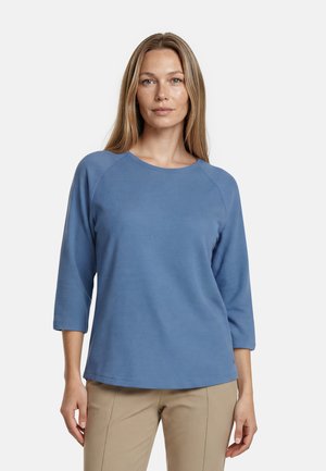 Femme aux longs cheveux raides portant un haut bleu à manches trois-quarts et un pantalon beige, debout devant un fond blanc.