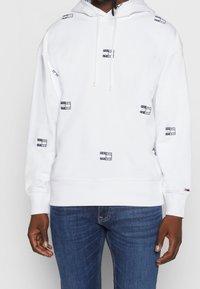 Witte hoodie met een capuchon met trekkoord, voorzien van een geborduurd logo patroon in marineblauw. Gemaakt van zachte stof, met geribbelde boorden aan mouwen en zoom.