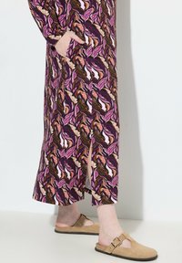 Vestido largo con un patrón de hojas púrpuras y rosas, hecho de una tela ligera. Presenta cortes laterales y bolsillos. Se lleva con sandalias de plataforma color beige.