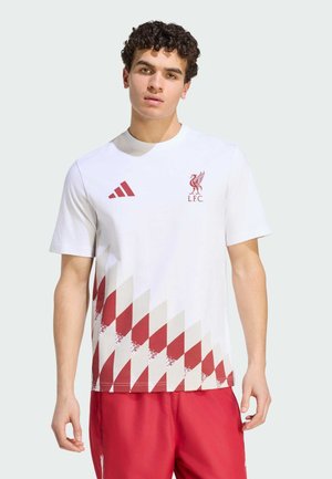 Młody mężczyzna w białej koszulce Liverpool FC z czerwonym geometrycznym wzorem i czerwonych szortach, stojący na jednolitym tle.