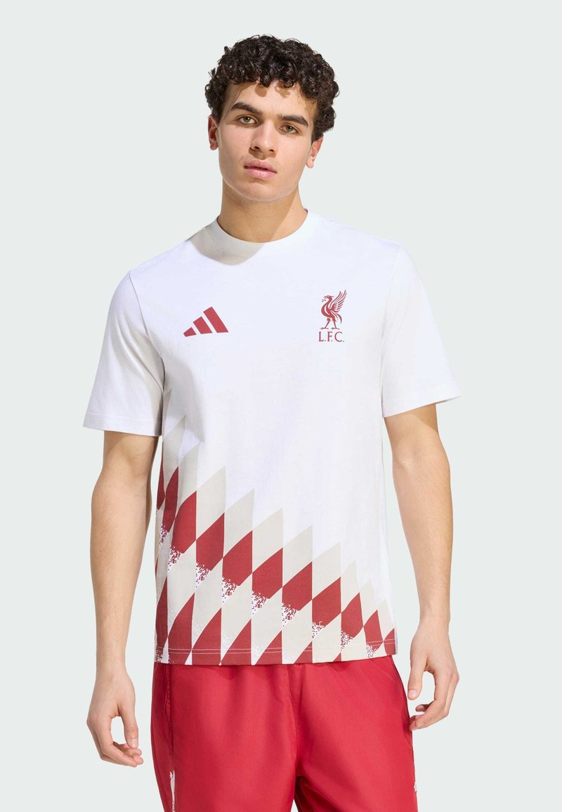 Jeune homme portant un t-shirt blanc du Liverpool FC avec un motif géométrique rouge et un short rouge, debout devant un fond uni.