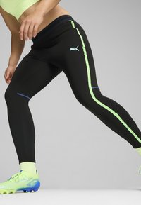 Fekete sportleggings, amely szoros illeszkedésű, neon zöld oldalsávokkal és kék díszítéssel a mandzsettákon. Az anyag rugalmasnak és simának tűnik.