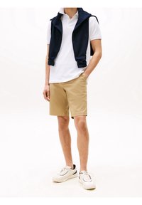 Mannenoutfit met een wit poloshirt, kaki shorts en een marineblauw mouwloos sweater dat over de schouders is gedrapeerd. Casual witte sneakers maken de look compleet.