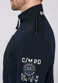 Felpa navy con zip caratterizzata da un alto colletto, tessuto a trama, toppe bianche e blu, e strisce a contrasto sulle spalle.