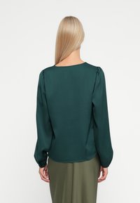 Bluza s dugim rukavima u tamnozelenom satenu s glatkom teksturom, okruglim izrezom i nabranim detaljima na ramenima. Uparena s maslinastom suknjom.