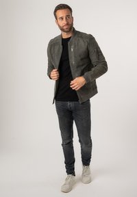 Giacca bomber in suede grigia con zip frontale e dettagli delle tasche, indossata sopra una maglietta nera, abbinata a jeans blu e sneakers bianche.