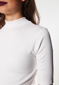 Hvit ribbet høyhalset topp med kort erme, med en tettsittende design og myk tekstur. Glatt finish, minimale hardware-detaljer.
