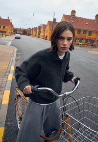 Ciemnoszary, oversize sweter z kołnierzem, połączony z jasnoszarymi spodniami dresowymi. Rower z metalowym koszykiem i kierownicą. W tle ulica i domy.