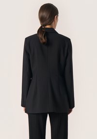 Schwarzer taillierter Blazer mit einem Rückenschlitz, Revers mit Kerbe und langen Ärmeln, kombiniert mit schwarzen, gerippten High-Waist-Hosen.