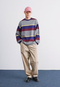 Pelēka garām piedurknēm krekls ar sarkaniem, ziliem un dzelteniem horizontāliem svītrām, bēša bikses un gaiši rozā cepure, apvienota ar melnām kurpēm.