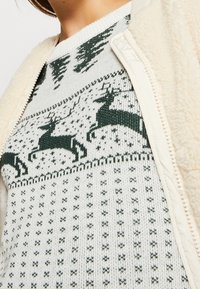Maglione a maglia bianco con motivi di cervi verdi, dotato di collo rotondo. Una giacca con zip color crema e texture è parzialmente visibile.