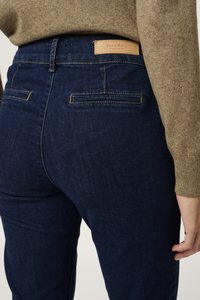 Sötétkék farmernadrág magas derekú fazonnal, két hátsó zsebbel és egy barna márkacímkével a hátsó derékrészen. Sima tapintású.