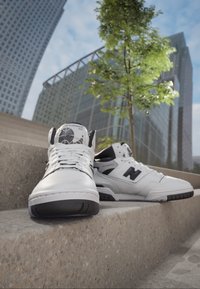 Białe skórzane buty sportowe za kostkę z czarnymi akcentami, wyposażone w teksturowaną podeszwę i detal z logo. Umieszczone na betonowych schodach.