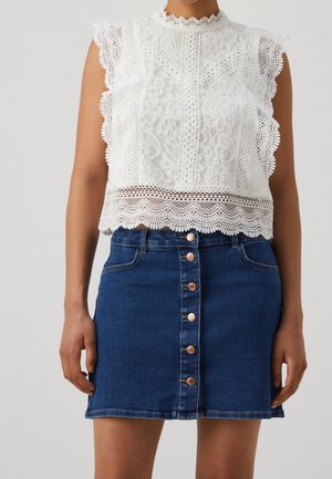 Woman wearing sleeveless white lace blouse with scalloped edges and high neck, paired with button-up dark blue denim mini skirt.