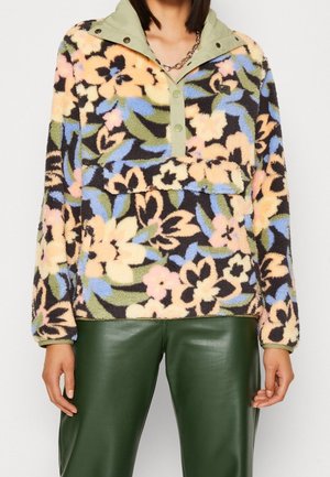 Frau trägt einen mehrfarbigen floralen Fleece-Pullover mit grünem Stehkragen mit Druckknöpfen und dunkelgrüner Lederhose, steht vor einem weißen Hintergrund.