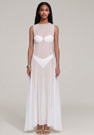 Donna che indossa un abito bianco trasparente senza maniche con biancheria intima bianca visibile e sandali dorati su uno sfondo chiaro uniforme.