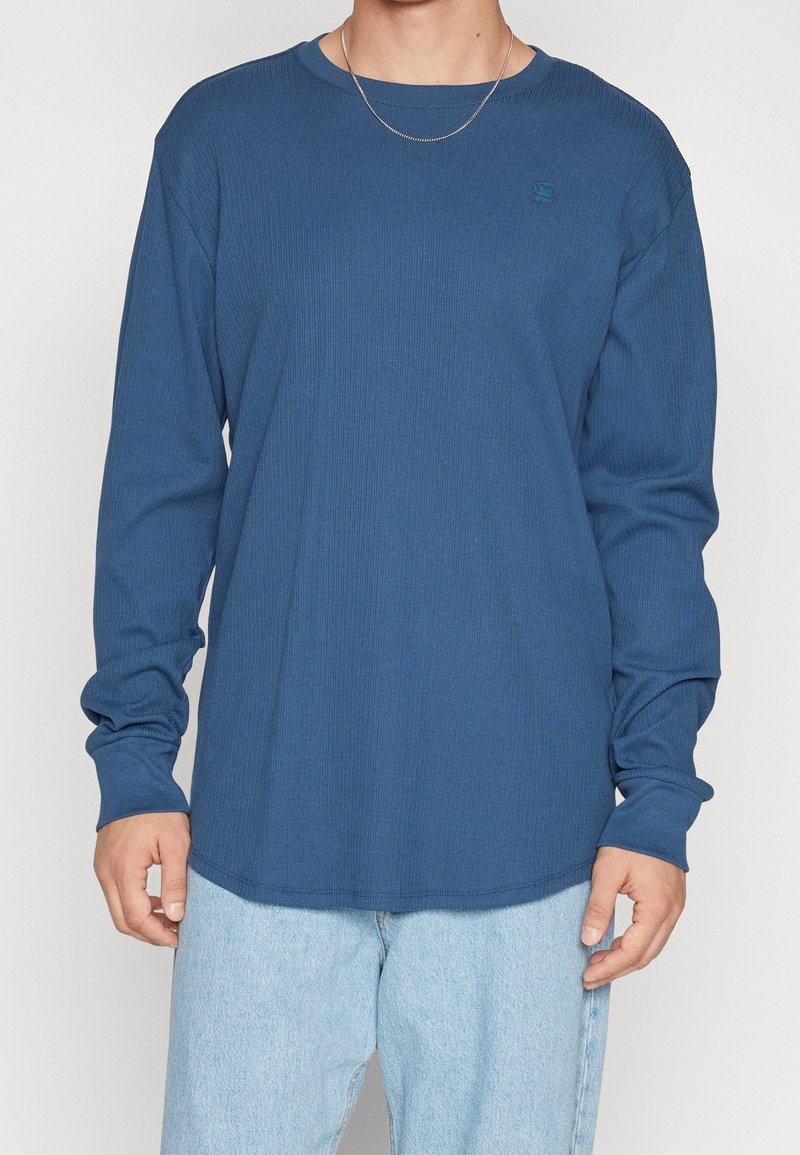 Personne portant une chemise bleue à manches longues et un jean bleu clair, avec une fine chaîne en argent, sur un fond uni.