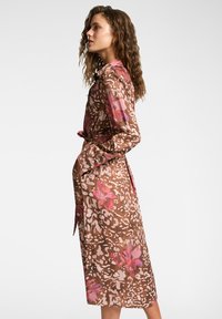 Trench coat marrone con motivi floreali rosa e bianchi, maniche lunghe, vita con cintura e design lungo fino al ginocchio, realizzato in tessuto leggero.