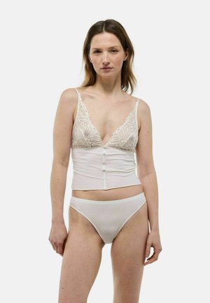 Femme à la peau claire et aux cheveux bruns portant un bralette en dentelle blanche et un sous-vêtement blanc côtelé assorti, debout devant un fond blanc.