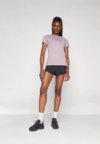 Γυναίκα που στέκεται με αυτοπεποίθηση φορώντας ένα ανοιχτό μωβ μπλουζάκι, μαύρες αθλητικές βερμούδες, λευκές κάλτσες και μαύρα αθλητικά παπούτσια, μπροστά σε έναν απλό φόντο.