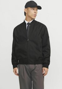 Chaqueta bomber negra con cierre de cremallera, puños y dobladillo de canalé. Acompañada de una gorra negra y una camisa formal con corbata. Pantalones grises.
