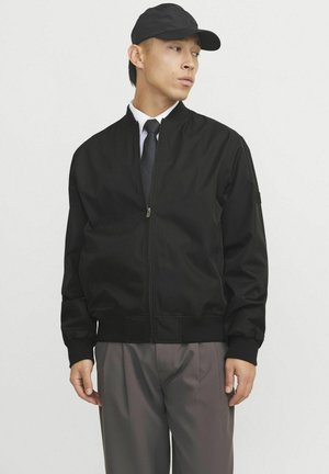 Schwarze Bomberjacke mit Reißverschluss, gerippten Bündchen und Saum. Begleitet von einer schwarzen Mütze und einem formellen Hemd mit Krawatte. Graue Hose.