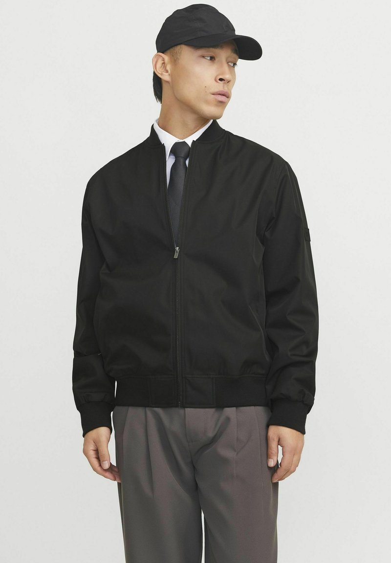 Chaqueta bomber negra con cierre de cremallera, puños y dobladillo de canalé. Acompañada de una gorra negra y una camisa formal con corbata. Pantalones grises.