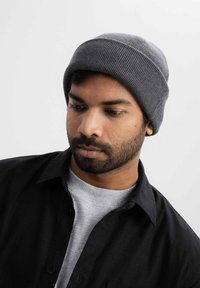 Grijze geribbelde gebreide beanie gedragen op zwart haar, gecombineerd met een zwart overhemd boven een grijze ronde halstrui. Neutrale achtergrond.