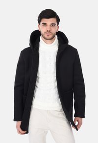 Cappotto nero con cappuccio, dalla texture liscia e due tasche frontali, indossato sopra un maglione bianco a trecce. Il modello è in piedi contro uno sfondo neutro.