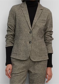 Texturerad houndsstooth-blazer i mörkbeige och svart, med två knappar, notched lapels och sidofickor, som bärs tillsammans med en svart polotröja.