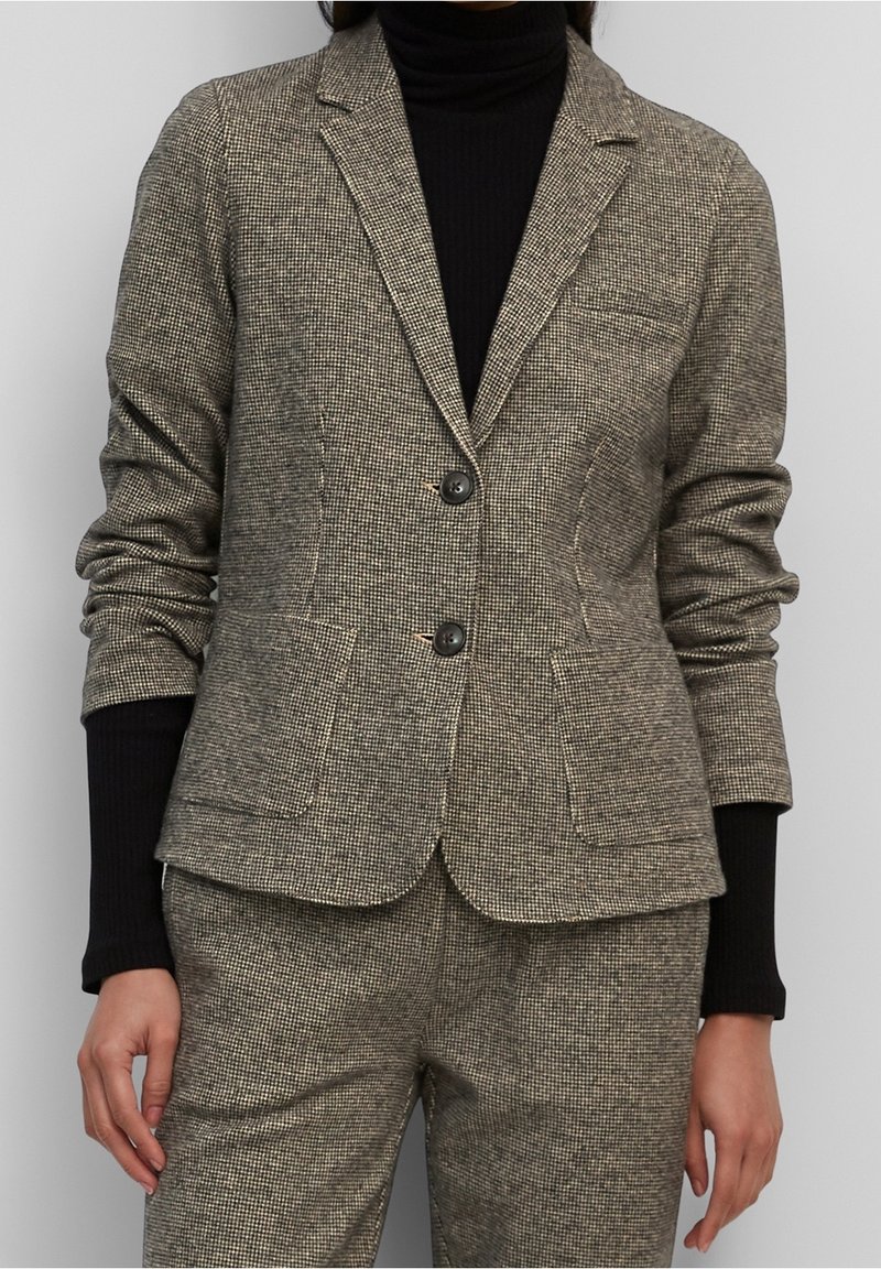 Texturerad houndsstooth-blazer i mörkbeige och svart, med två knappar, notched lapels och sidofickor, som bärs tillsammans med en svart polotröja.
