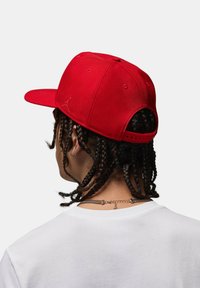 Rode baseballpet met een vlakke rand, verstelbare achterkant en een klein logo aan de zijkant. Gedragen door een persoon met gevlochten haar en een wit T-shirt.